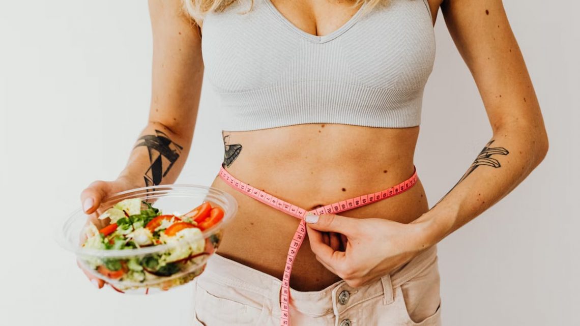 Woman measuring waistline while holding a fresh vegetable salad, emphasizing healthy living.