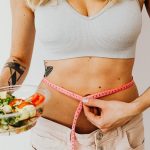 Woman measuring waistline while holding a fresh vegetable salad, emphasizing healthy living.