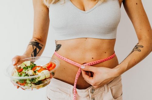 Woman measuring waistline while holding a fresh vegetable salad, emphasizing healthy living.